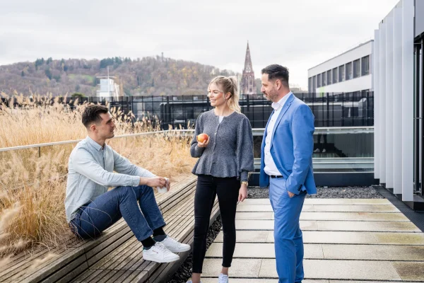 Drei Personen befinden sich auf einer Terrassen in einem Austausch, wobei eine Person auf einer Bank sitzt und zwei weitere Personen davor stehen